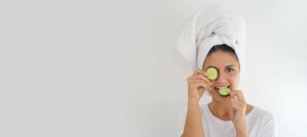 Vibrant brunette with towel turban and cucumber on eye on white background - Photo, Image
