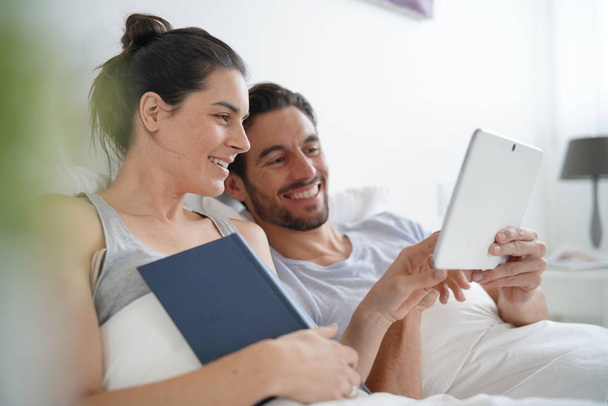 Attractive couple reading and relaxing in bed with book and tablet - Foto, immagini