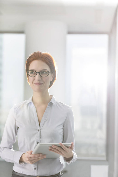 Улыбающаяся молодая деловая женщина, использующая цифровой планшет, стоя в офисе
 - Фото, изображение
