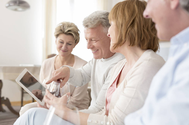 Sorrindo homem maduro segurando tablet digital enquanto sentado com amigos em casa
 - Foto, Imagem