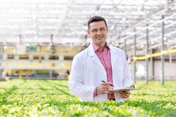 Portrait of smiling scientist standing with clipboard amidst herbs in greenhouse - Foto, imagen