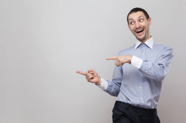 Portrait of funny businessman pointing at side copyspace and looking aside with amazed face on grey background - Foto, afbeelding