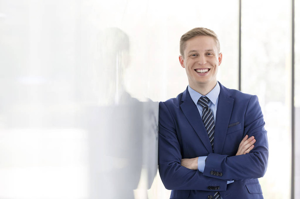 Portrait of smiling young businessman with arms crossed leaning on wall at office - Fotó, kép