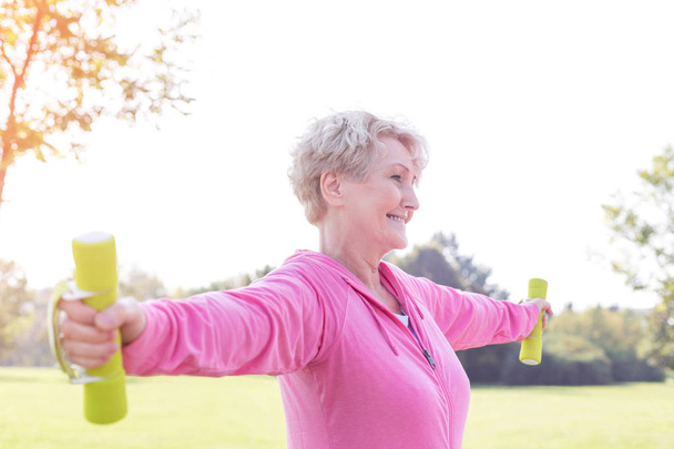 Senior woman with arms outstretched lifting dumbbells in park - Foto, afbeelding