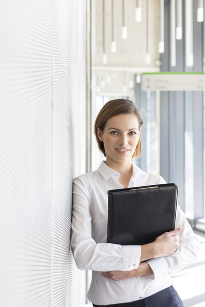 Portrait of businesswoman holding file by wall at office - Fotoğraf, Görsel
