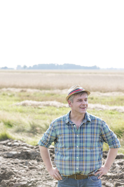 Glimlachend volwassen boer in geruite hemd staan met handen op de heupen op boerderij - Foto, afbeelding