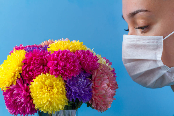 Allergy concept. Allergy to flowers bloom and pollen. A young woman in a medical mask and flowers on a blue background. Allergens, allergic reaction - Fotografie, Obrázek