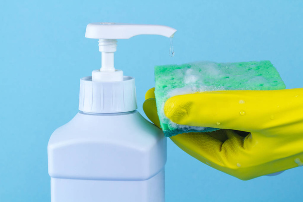 Dishwashing detergent and hand in yellow gloves with a dishwashing sponge on a blue background. Cleaning the kitchen, washing dishes - Фото, зображення