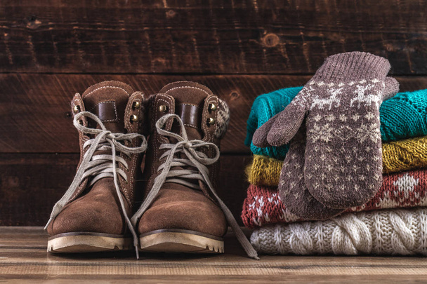 Knitted, winter, folded sweaters, warm mittens and winter boots on a wooden background. Winter clothes. Ugly Christmas Sweater. Warm, comfortable clothes - Foto, Imagen