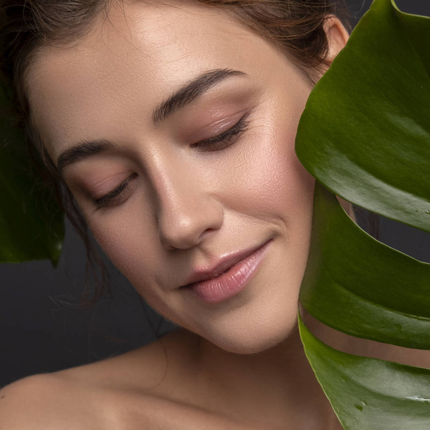 Cute ordinary young woman posing with monstera tropical leafs. Fresh clean skin with flawless texture. Skin care beauty cosmetology concept. Space for text. - Photo, Image