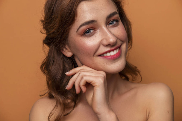 Cute caucasian brunette woman with curly hairstyle on orange background. Fresh clean skin with evening make up and freckles. Hands near her face. Close up portrait. - Photo, Image