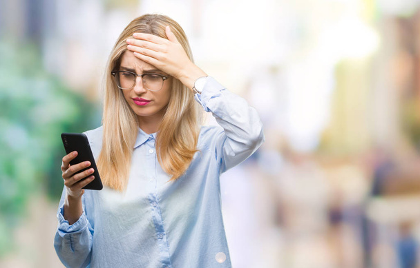 Young beautiful blonde business woman using smartphone over isolated background stressed with hand on head, shocked with shame and surprise face, angry and frustrated. Fear and upset for mistake. - Photo, Image
