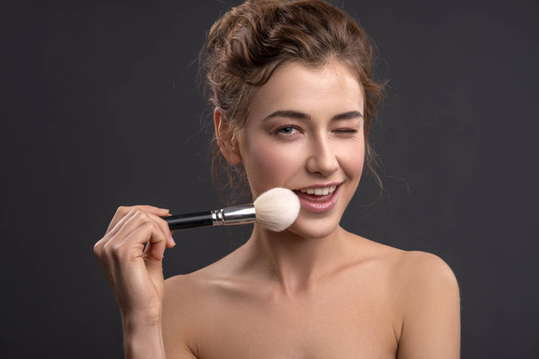 Beautiful brunette caucasian young woman prepare herself, applying powder on her cheeks with a brush. Clean, fresh, natural, flawless skin. Soft smile on her face. Close up on a neutral background - Photo, Image