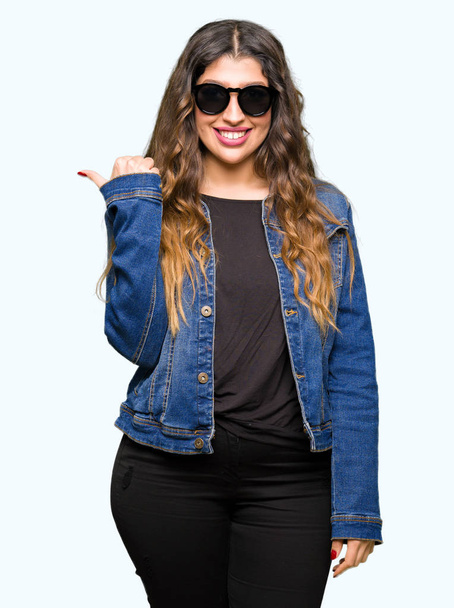 Young beautiful woman wearing sunglasses smiling with happy face looking and pointing to the side with thumb up. - Foto, Imagen