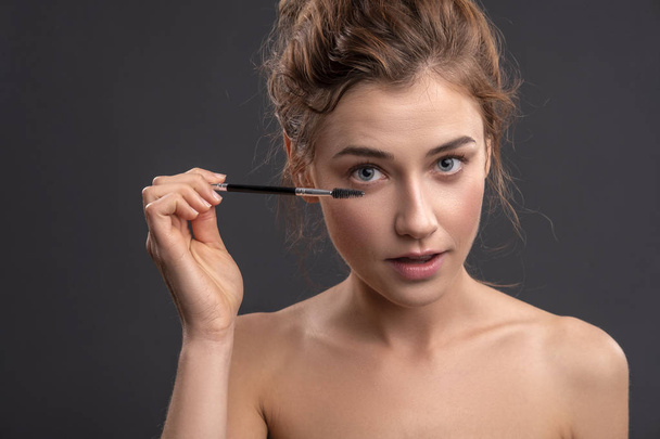 Beautiful brunette caucasian young woman prepare herself, applying mascara on her eyes with a brush. Clean, fresh, natural, flawless skin. Soft smile on her face. Close up on a neutral background - 写真・画像
