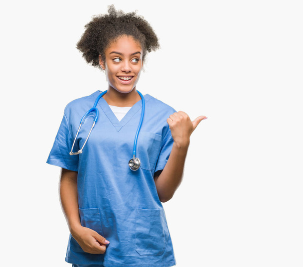 Young afro american doctor woman over isolated background smiling with happy face looking and pointing to the side with thumb up. - Fotografie, Obrázek