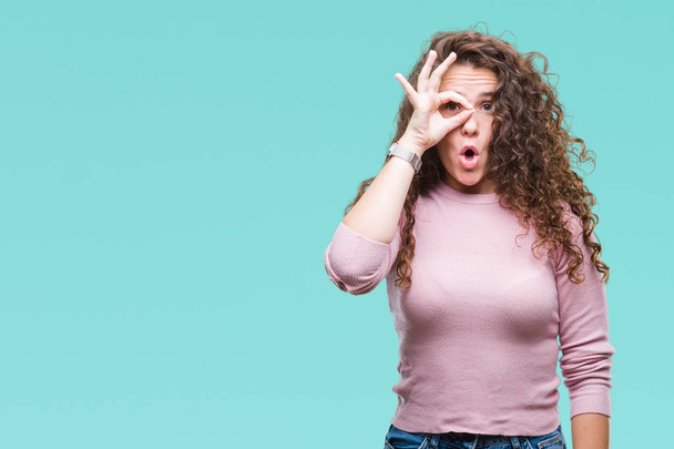 Beautiful brunette curly hair young girl wearing pink sweater over isolated background doing ok gesture shocked with surprised face, eye looking through fingers. Unbelieving expression. - Photo, Image