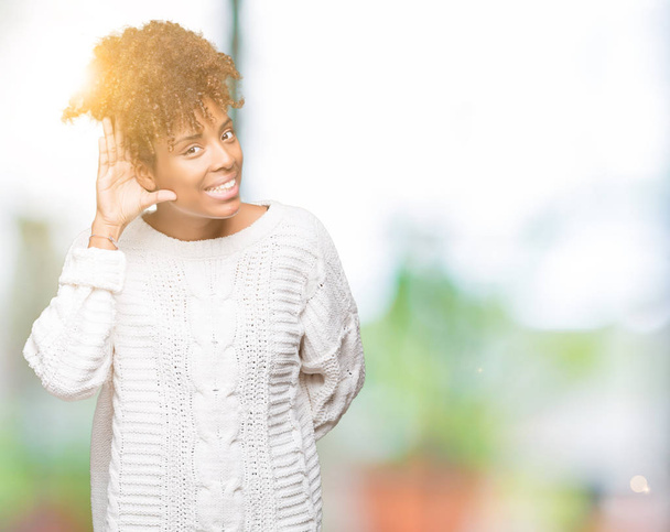 Belle jeune femme afro-américaine portant un pull d'hiver sur fond isolé souriant avec la main sur l'oreille en écoutant une rumeur ou des commérages. Concept de surdité
. - Photo, image