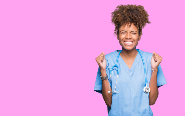 Молодая африканская американка-врач на изолированном фоне, взволнованная успехом с поднятыми руками, радовалась победе, улыбаясь. Концепция победителя
. - Фото, изображение