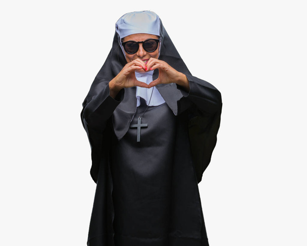 Middle age senior catholic nun woman wearing sunglasses over isolated background smiling in love showing heart symbol and shape with hands. Romantic concept. - Photo, Image