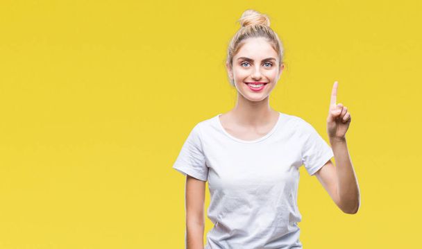 Young beautiful blonde woman wearing white t-shirt over isolated background showing and pointing up with finger number one while smiling confident and happy. - Photo, Image