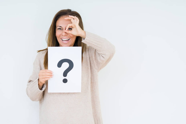 Middle age woman thinking and holding paper with question mark symbol over isolated background with happy face smiling doing ok sign with hand on eye looking through fingers - Foto, Imagem