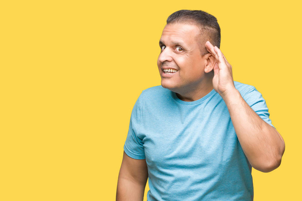 Uomo arabo di mezza età che indossa t-shirt blu su sfondo isolato sorridente con mano sull'orecchio ascoltando un'udienza a pettegolezzi o pettegolezzi. Concetto di sordità
. - Foto, immagini