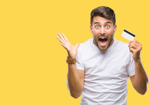 Young handsome man holding credit card over isolated background very happy and excited, winner expression celebrating victory screaming with big smile and raised hands - Photo, Image