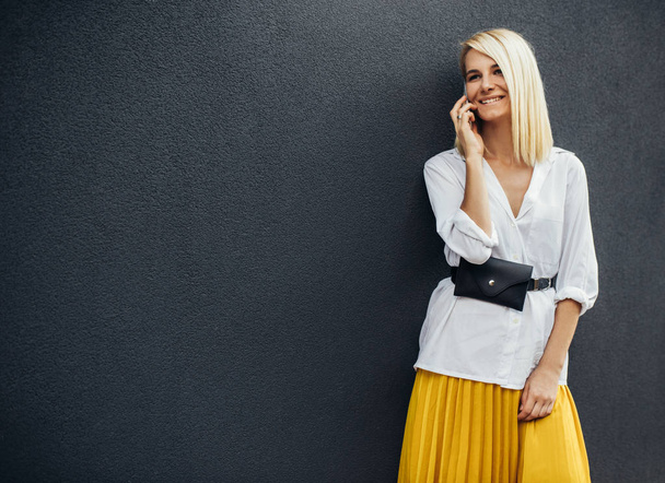 blonde woman smiling and standing against gray wall while talking on smart phone - Foto, Imagen