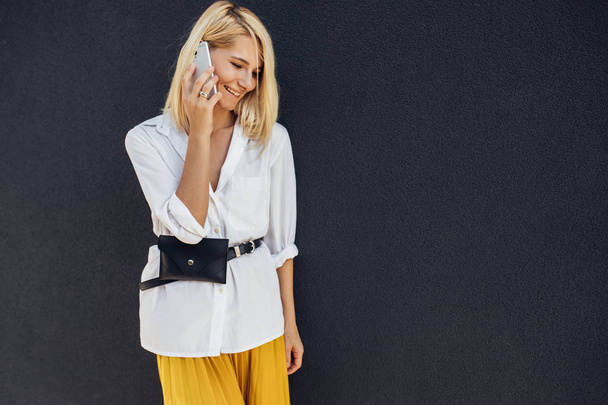 blonde woman smiling and standing against gray buildings wall while talking on mobile phone - Fotó, kép