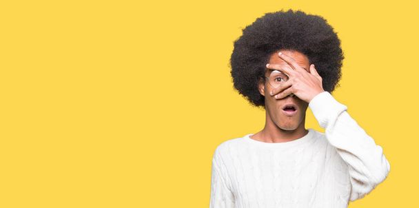 Jeune homme afro-américain aux cheveux afro portant des lunettes jetant un coup d "œil sur le visage et les yeux avec la main, regardant à travers les doigts avec une expression embarrassée
. - Photo, image