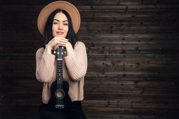 Jong meisje glimlachend met een ukulele zitten in studio op een houten achtergrond. - Foto, afbeelding