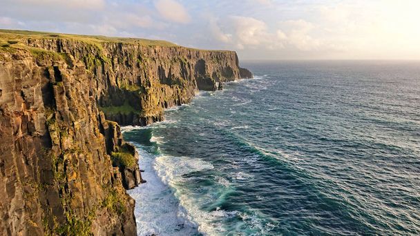 Írország nyugati partja panoráma. Az előtérben látható a sziklák és sziklák, a madarak, a fű, a tehenek, a hullámok, a spray. A háttérben van a tenger, a felhők és a sirályok. Moher-sziklák - Fotó, kép
