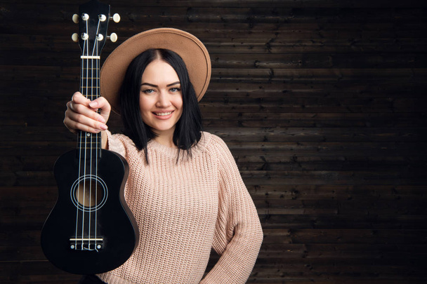 Young funny hipster girl having fun and playing on small ukulele guitar, singing and dancing. wearing vintage hat, joy, positive mood. Dark wooden wall background. - Foto, imagen