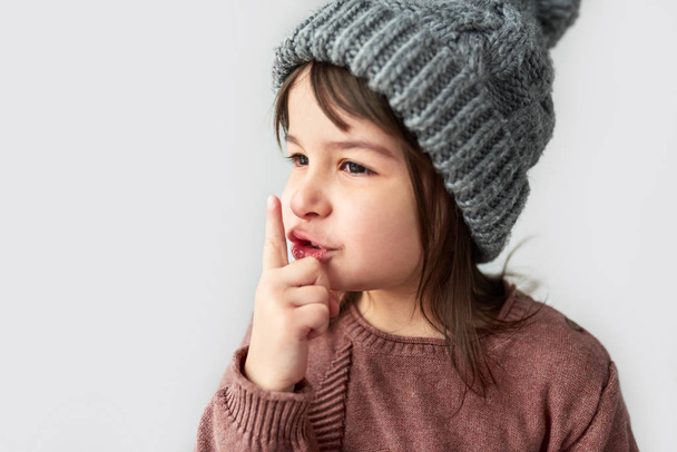 cute little girl in winter warm gray hat, wearing sweater and showing silent gesture - 写真・画像