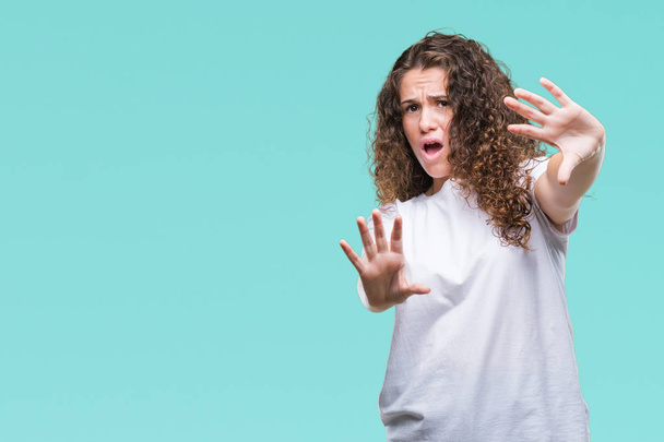 Beautiful brunette curly hair young girl wearing casual t-shirt over isolated background afraid and terrified with fear expression stop gesture with hands, shouting in shock. Panic concept. - Foto, Bild