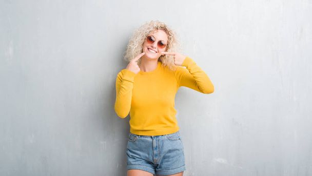 Jeune femme blonde aux cheveux bouclés sur fond gris grunge souriant confiant montrant et pointant avec les doigts les dents et la bouche. Concept de santé
. - Photo, image