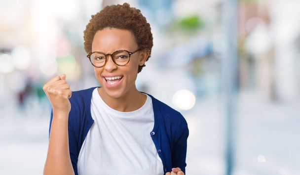 Young beautiful african american woman wearing glasses over isolated background very happy and excited doing winner gesture with arms raised, smiling and screaming for success. Celebration concept. - Photo, Image