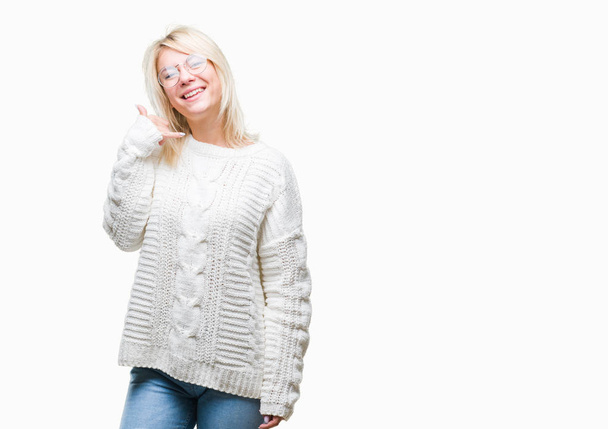 junge schöne blonde Frau mit Winterpullover und Brille vor isoliertem Hintergrund lächelt und macht Telefongesten mit Händen und Fingern, als spräche sie am Telefon. Vermittlung von Konzepten. - Foto, Bild