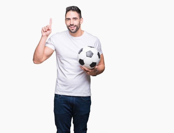 Młody człowiek posiadający piłka nożna Piłka nożna na białym tle zaskoczony idei lub pytanie palcem wskazującym z szczęśliwy twarz, numer jeden - Zdjęcie, obraz