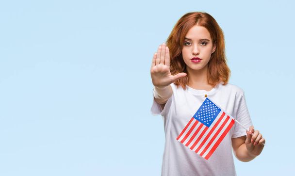 Jovem bela mulher segurando bandeira da América sobre fundo isolado com a mão aberta fazendo sinal de parada com expressão séria e confiante, gesto de defesa
 - Foto, Imagem