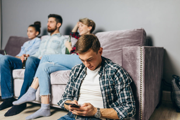 young man in checked shirt is sending sms while his friends are watching TV - 写真・画像