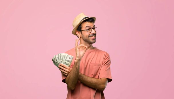 Man holding many bills showing ok sign while winking an eye on pink background - Zdjęcie, obraz
