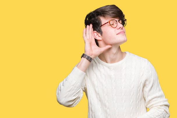 Mladý pohledný muž nosí brýle izolované pozadí s úsměvem s rukou přes ucho naslouchá slyšení zvěsti nebo drby. Hluchota koncept. - Fotografie, Obrázek