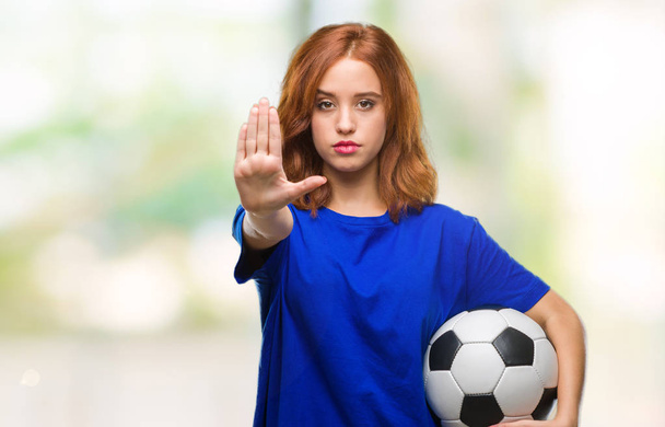 Молодая красивая женщина на изолированном фоне, держа футбольный мяч с открытой рукой делает знак стоп с серьезным и уверенным выражением, защитный жест
 - Фото, изображение