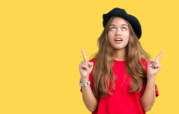 Young beautiful brunette fashion woman wearing red t-shirt and black beret over isolated background amazed and surprised looking up and pointing with fingers and raised arms. - Photo, Image