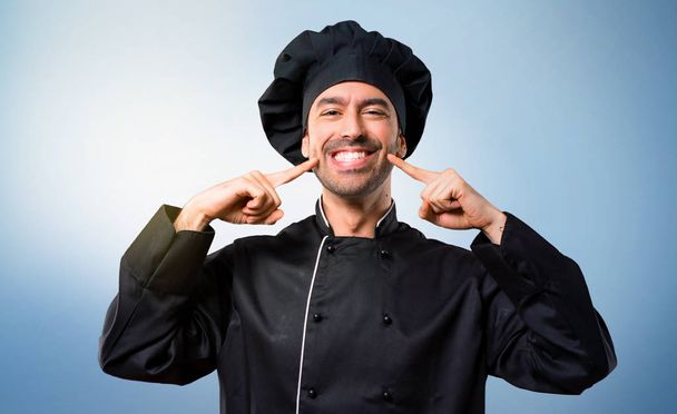 Chef-kok man In het zwart uniform glimlachend, met een gelukkig en aangename uitstraling terwijl mond en gezicht met vingers wijzen op blauwe achtergrond - Foto, afbeelding