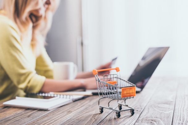 selective focus of small shopping trolley with woman using laptop on background  - Фото, зображення