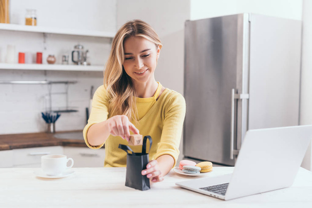 mulher atraente colocando pequena caixa de papel em saco de compras preto na cozinha
  - Foto, Imagem