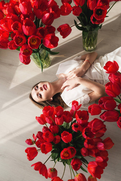 Una hermosa joven en un peignoir está tirada en el suelo entre grandes ramos de tulipanes. 8 de marzo concepto
. - Foto, imagen